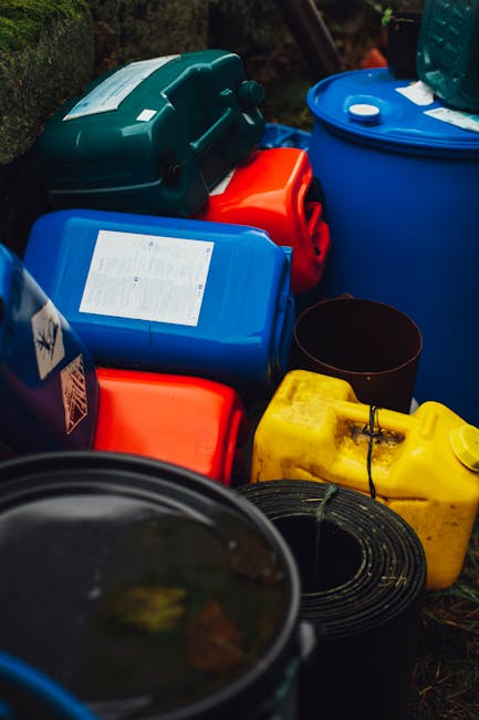 A collection of large plastic barrels with orange and black lids, arranged outdoors on a flat surface. The barrels vary in color, with some being white and others blue, all with secure lids. In the ba