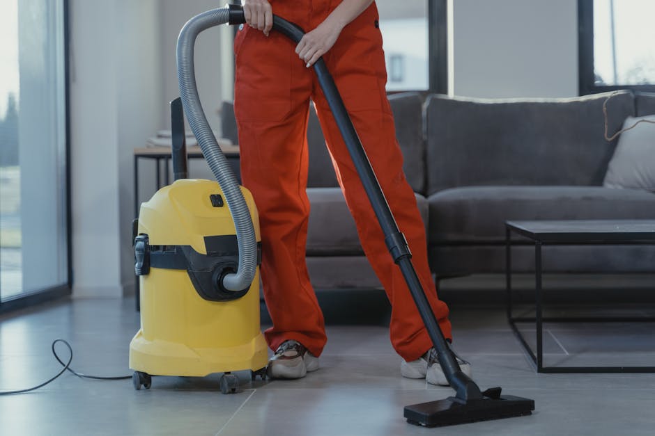 A person dressed in orange work trousers and white sneakers uses a yellow and black industrial vacuum cleaner placed on a gray tile floor within a modern living room. The individual holds the vacuum's