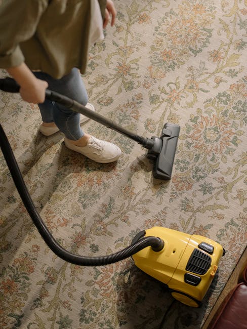 A professional in full protective PPE, including a white suit, face mask, gloves, and goggles, performing surface cleaning or sanitisation in a modern, well-lit living room. The individual is using a 