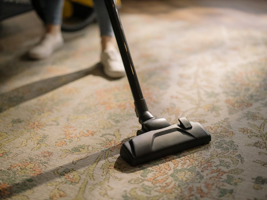 Close-up of a vacuum cleaner head with a black handle and a switch, actively cleaning a patterned area rug with floral motifs in shades of cream, orange, and green. The rug covers a wooden floor, and a person wearing white shoes and dark pants is partially visible in the background, indicating a domestic setting. The scene is well-lit, emphasizing the surface cleanliness and the deep cleaning process carried out by Carpet Cleaners Putney, exemplifying thorough surface cleaning and maintenance in a residential space.