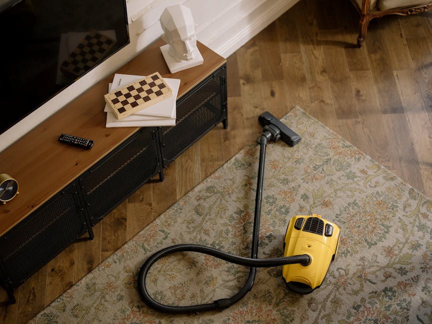 Close-up of a vacuum cleaner head with a black handle and a switch, actively cleaning a patterned area rug with floral motifs in shades of cream, orange, and green. The rug covers a wooden floor, and a person wearing white shoes and dark pants is partially visible in the background, indicating a domestic setting. The scene is well-lit, emphasizing the surface cleanliness and the deep cleaning process carried out by Carpet Cleaners Putney, exemplifying thorough surface cleaning and maintenance in a residential space.