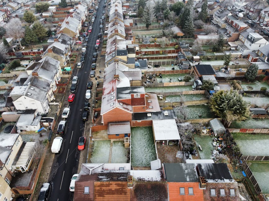 An aerial view of a residential neighborhood showing closely packed houses with pitched roofs, some with solar panels, surrounded by trees and gardens. The streets are lined with parked cars, and the 